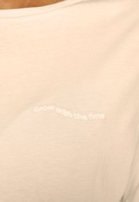 T-shirt bege claro feito de um tecido suave, com um bordado curvo a dizer "Cresça com o fluxo" em fio branco na parte frontal.