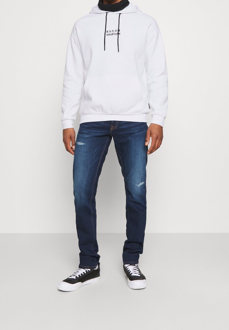 Homme portant un sweat à capuche blanc avec des cordons noirs et un texte, jean bleu foncé légèrement usé, et baskets noires avec semelles blanches.