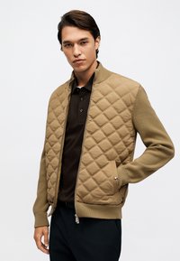 Άνδρας που φορά ένα καφέ μπουφάν με καπιτονέ ύφασμα και πλεκτά μανίκια πάνω από μια μαύρη πόλο μπλούζα, στέκεται με το ένα χέρι στην τσέπη του μπροστά σε λευκό φόντο.