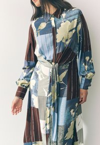 Bunt gestreiftes Kleid mit Kragen, langen Ärmeln, floralen Mustern und einer gebundenen Taille. Hergestellt aus einem glatten, leichten Stoff mit verschiedenen Texturen.