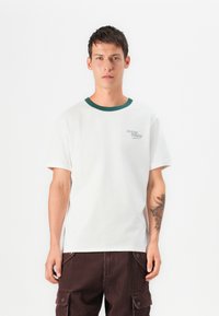 Camiseta de algodón blanca con cuello acanalado verde, que presenta un pequeño logotipo de "Cowboy Country" en el lado izquierdo del pecho. El modelo lleva pantalones de pana marrón oscuro.