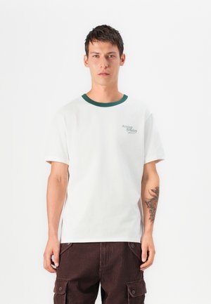Camiseta de algodón blanca con cuello acanalado verde, que presenta un pequeño logotipo de "Cowboy Country" en el lado izquierdo del pecho. El modelo lleva pantalones de pana marrón oscuro.
