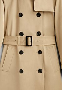 Trench-coat beige croisé avec boutons noirs, ceinture avec boucle rectangulaire et deux poches latérales.