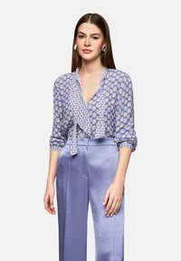 Himmelblaue Bluse mit V-Ausschnitt, Taillenknoten und gemusterter Design; kombiniert mit hochtaillierten, hellvioletten, weiten Hosen.