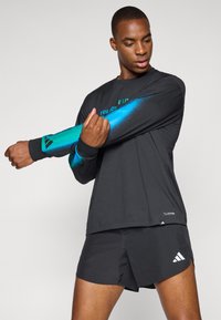 Črna dolgosleeve atletska majica z akcenti v teal gradaciji in zelenim napisom, v kombinaciji s črnimi kratkimi hlačami z belimi logotipnimi detajli.