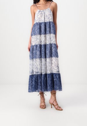 Kobieta w bezrękawną, warstwową suknię maxi z niebiesko-białym kwiatowym wzorem oraz cielistymi sandałkami na wysokim obcasie ze sznurowaniem, stojąca na białej podłodze.