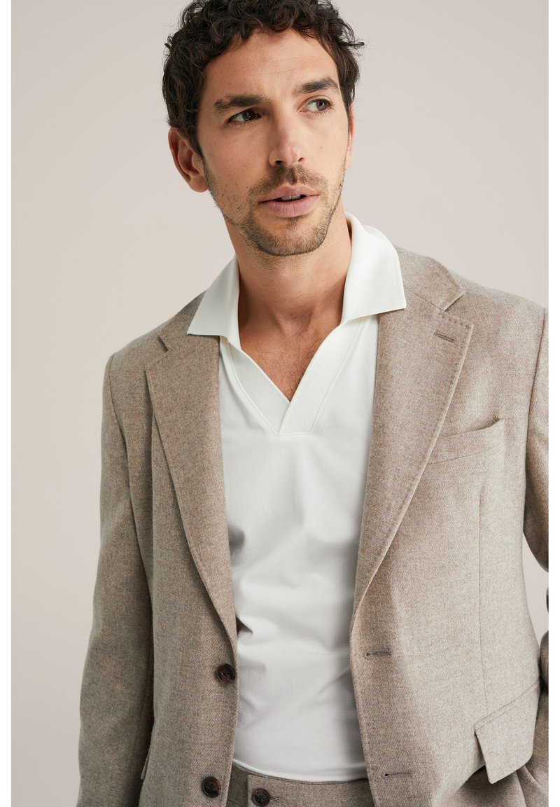 Homme aux cheveux courts et foncés portant un blazer beige et un t-shirt blanc à col en V, regardant légèrement vers sa gauche sur un fond neutre.