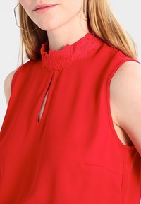 Červená halenka bez rukávů s texturovaným krajkovým límcem, průstřihem na hrudi a malou kapsou na boku, vyrobená z hladké látky.