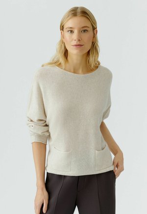 Pull en tricot beige clair avec un col rond, coupe décontractée et deux poches avant. Tissu texturé et manches légèrement roulées.