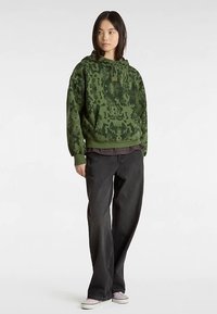 Zielona wzorzysta bluza z kapturem z miękkiego materiału, z przednią kieszenią i regulowanym kapturem na sznurek, noszona z ciemnymi dżinsami i jasnymi trampkami.