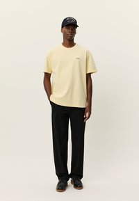 Ανοιχτό κίτρινο βαμβακερό μπλουζάκι με κοντά μανίκια, συνδυασμένο με μαύρο παντελόνι και μαύρα παπούτσια. Μια σκούρα καπέλο με λογότυπο ολοκληρώνει την εμφάνιση.