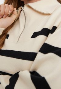 Beżowy sweter z czarnymi poziomymi paskami, z kołnierzem zapinanym na zamek i miękką teksturą. Zbliżenie na materiał i detale zamka.