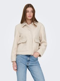 Veste courte couleur crème en tissu texturé avec col pointu, deux poches avant et fermeture à boutons, associée à un jean bleu.