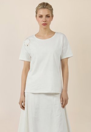 Blonde Frau mit zurückgebundenem Haar trägt ein schlichtes weißes kurzärmeliges Oberteil mit Ausschnittdetail und einen weißen A-Linien-Rock vor beigem Hintergrund.