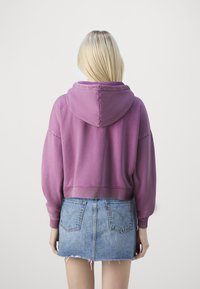 Sudadera corta morada con capucha y mangas anchas, combinada con una minifalda vaquera desgastada que tiene bolsillos traseros.