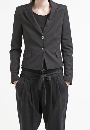 Schwarzer taillierter Blazer mit zwei Knöpfen über einem schwarzen Hemd, kombiniert mit locker sitzenden schwarzen Hosen, die an der Taille gebunden sind, Hände in den Taschen.