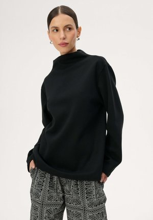 Kobieta o ciemnych włosach związanych w niski kok, nosząca srebrne kolczyki koła, czarną bluzkę z długim rękawem oraz czarno-białe spodnie w wzór paisley.