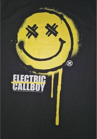 Svart t-shirt med en stor gul smiley-ansikte grafik med korslagda linjer som ögon, droppande målar effekt, och vit text "ELECTRIC CALLBOY."