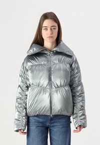 Casaco puffer prateado metálico com um grande colarinho, fecho de correr frontal, detalhes elásticos e um corte solto. Usado sobre jeans azuis.