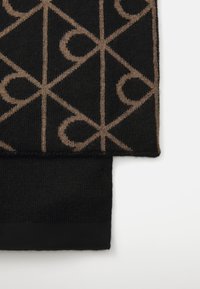 Nærbilde av foldet svart strikket stoff med beige geometrisk mønster og solid svart ribbet kant på hvit bakgrunn.