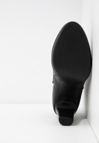 Botte noire à talon positionnée debout avec la semelle face vers l'avant, sur un fond blanc uni avec des coins.