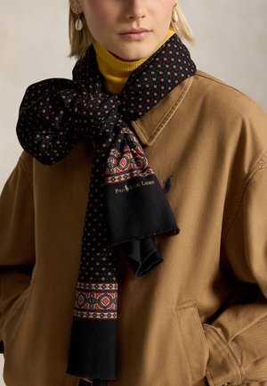 Vrouw draagt een beige jas en een gele coltrui met een zwart patroon van een Polo Ralph Lauren sjaal, gebonden in een grote strik rond haar nek.