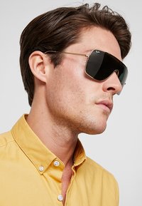 Occhiali da sole con lenti nere, montatura in metallo dorato e forma panoramica. Indossati da una persona con una camicia gialla a bottoni.