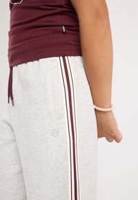 Vue latérale d'une personne portant un pantalon de survêtement gris avec des rayures bordeaux et blanches et un t-shirt bordeaux, la main relâchée sur le côté avec un bracelet.
