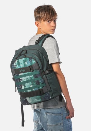 Ragazzo con t-shirt grigia e jeans guarda indietro mentre porta un grande zaino verde e nero con fantasia e molte cinghie.