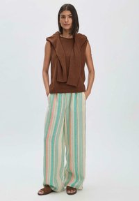 Bruine mouwloze trui met een kap, gecombineerd met brede gestreepte broeken in muntgroen, crème en pastelroze, en bruine sandalen.