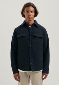 Młody mężczyzna z średniej długości kręconymi włosami, ubrany w ciemnoniebieską koszulę z kieszeniami na klatce piersiowej i beżowe spodnie.