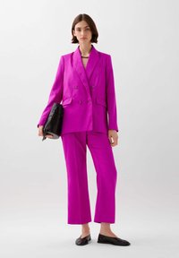 Blazer fucsia de doble botonadura con un patrón texturizado, combinado con pantalones a juego. La modelo sostiene un bolso negro y lleva zapatos planos negros.