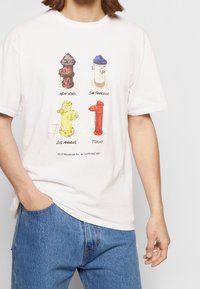 Vit bomullst-shirt med illustrationer av fyra brandposter i brunt, vitt, gult och rött, märkta med stadens namn.