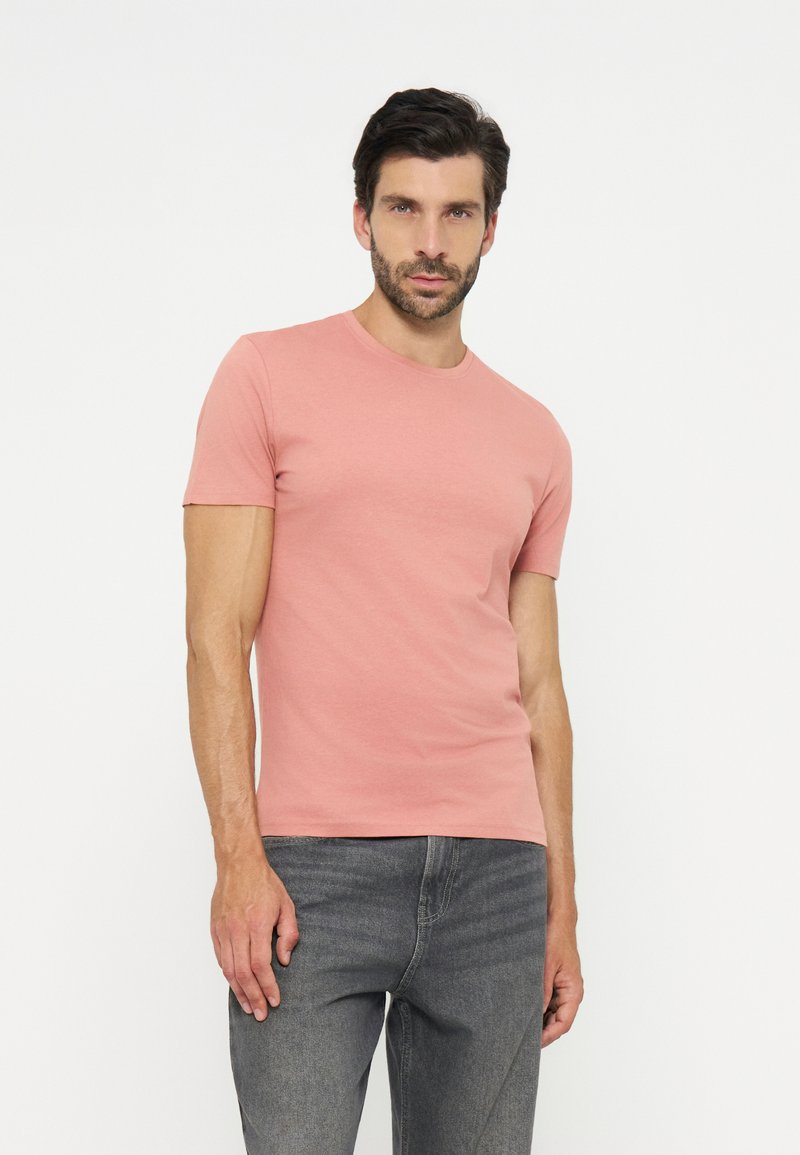 Man met donker haar en baard, die een aansluitend roze T-shirt en grijze spijkerbroek draagt, staand tegen een effen witte achtergrond.