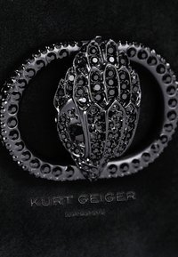 Fibbia a forma di mano con perline nere, con dettagli intricati, su uno sfondo nero che presenta il testo in rilievo "Kurt Geiger London".