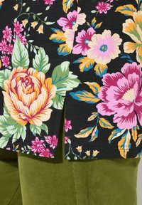 Bluse mit floralem Muster aus schwarzem Stoff, versehen mit großen Rosen, Pfingstrosen und Grünpflanzen; kurzer Schlitz am Saum, kombiniert mit grünen Cordhosen.