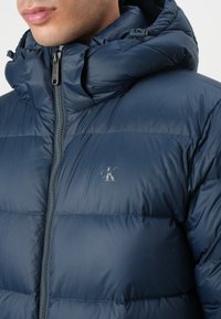 Navy blue puffer jacket with a zippered front, high collar, and hood. Features quilted sections and a small embossed logo on the chest.