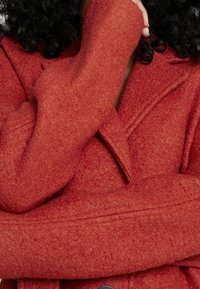 Czerwony wełniany płaszcz o teksturowanej tkaninie, złożonymi rękawami i z wydłużonym kołnierzem. Posiada ukryte zapięcie na guziki oraz minimalne detale przeszycia.