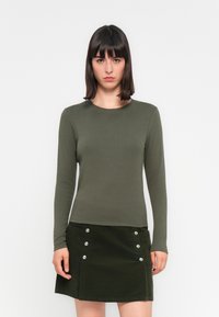 Haut à manches longues, olive fabriqué en tissu lisse, associé à une jupe en velours côtelé vert foncé, dotée de boutons-pression en argent et de poches.
