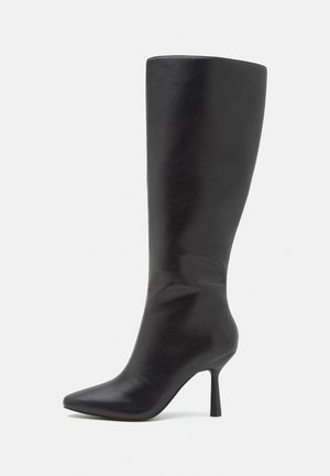 Botte en cuir noire montant jusqu'au genou, avec bout pointu et talon haut fin, sur fond blanc.