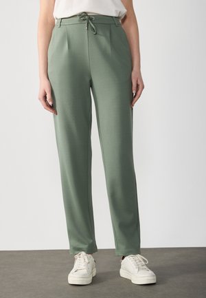 Personne portant un pantalon droit vert taille haute avec un cordon à la taille et des baskets blanches, debout sur un sol gris.