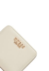Portafoglio sintetico beige con logo inciso 'GUESS EST 1981', texture liscia, angoli arrotondati e chiusura con zip argentata.