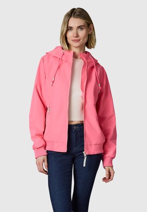 Femme portant une veste à capuche rose vif par-dessus un pull court blanc et un jean bleu foncé, debout devant un fond clair uni.