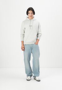 Felpa grigia chiaro con cappuccio e logo sul davanti, abbinata a jeans azzurri a vestibilità ampia e sneakers bianche con dettagli neri.