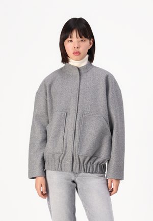 Veste en mélange de laine grise avec un col montant, des épaules tombantes et un ourlet élastique. Dispose de deux grandes poches avant et d'une texture douce.