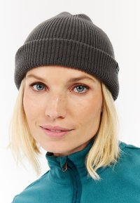 Gebreide grijze beanie met een geribbelde textuur, strakke pasvorm en een klein logolabel aan de zijkant, gecombineerd met een teal fleecejack met hoge kraag.