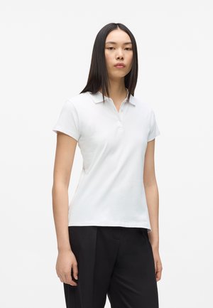 Ung kvinde med glat sort hår iført en hvid kortærmet poloshirt og sorte bukser, stående mod en ensfarvet lys baggrund.