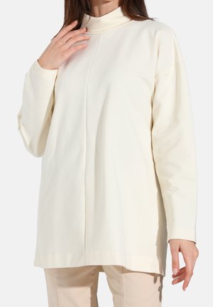 Femme portant un haut tunique crème à manches longues et col montant avec une couture centrale devant, associée à un pantalon beige clair.