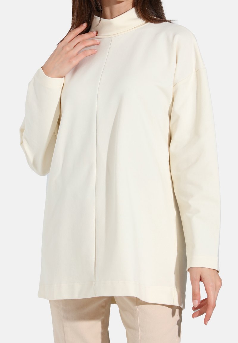 Femme portant un haut tunique crème à manches longues et col montant avec une couture centrale devant, associée à un pantalon beige clair.