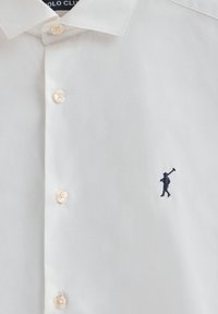 Camisa blanca de botones con tres botones visibles y un pequeño logotipo de jugador de polo en azul marino bordado en el lado derecho.